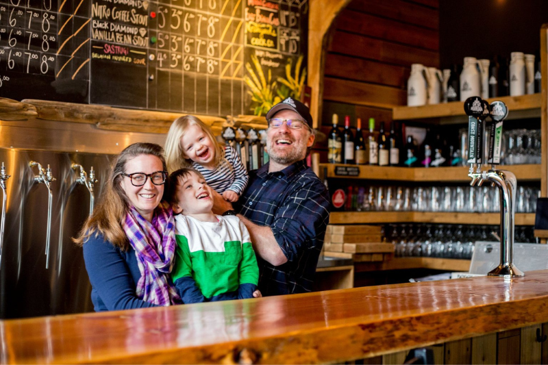 Family of four laughing and smiling standing behind a bar in a brewery (Coast Mountain Brewing) - Whiski Jack Resorts Whistler