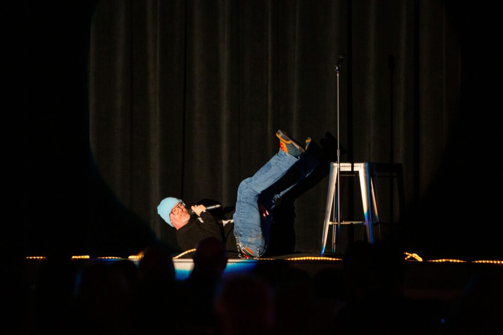 A comedian on stage laying down telling a joke next to a stool and a microphone stand