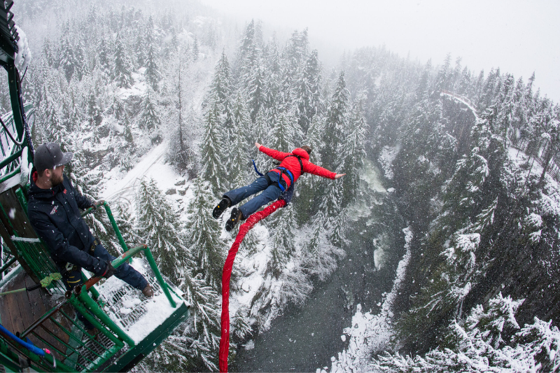 person bungee jumping over a river surrounded by snow, wearing a red jacket, at Whistler Bungee.