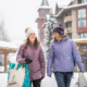 Two women walking outside in the snow in whistler village wearing winter clothing, carrying shopping bags, laughing.